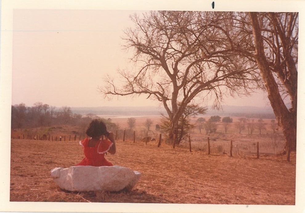 girl in red dress looking out to dry savannah through binoculars. dry trees on right of girl on white rock and clear sky.